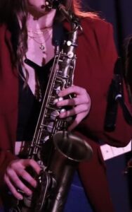 Image of a young woman in a maroon blazer, playing the saxaphone. She has long brown hair and multiple gold chains around her neck.